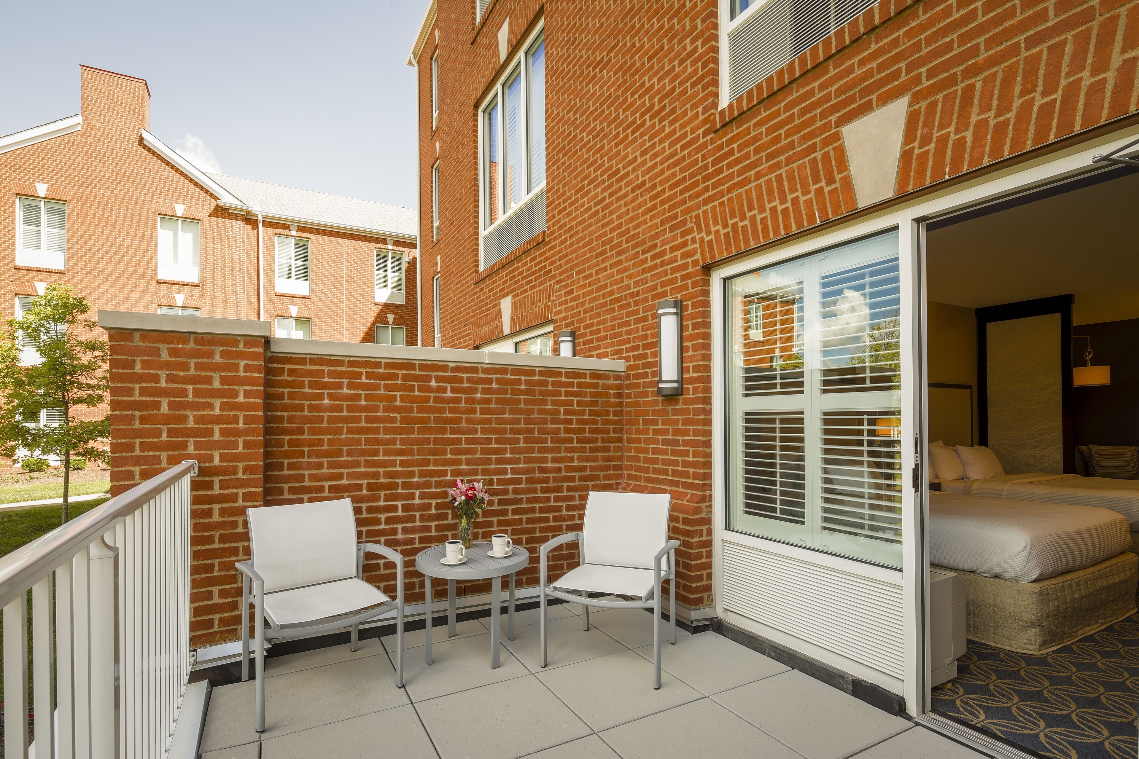 Un espace patio avec deux chaises blanches et une petite table, une porte-fenêtre coulissante donnant sur une chambre, et un bâtiment en briques en arrière-plan.