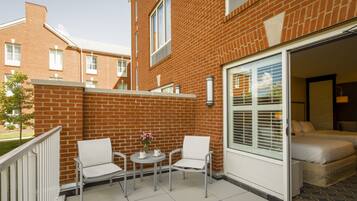 Un espace patio avec deux chaises blanches et une petite table, une porte-fenêtre coulissante donnant sur une chambre, et un bâtiment en briques en arrière-plan.