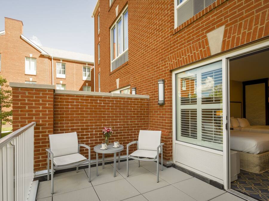 Un espace patio avec deux chaises blanches et une petite table, une porte-fenêtre coulissante donnant sur une chambre, et un bâtiment en briques en arrière-plan.