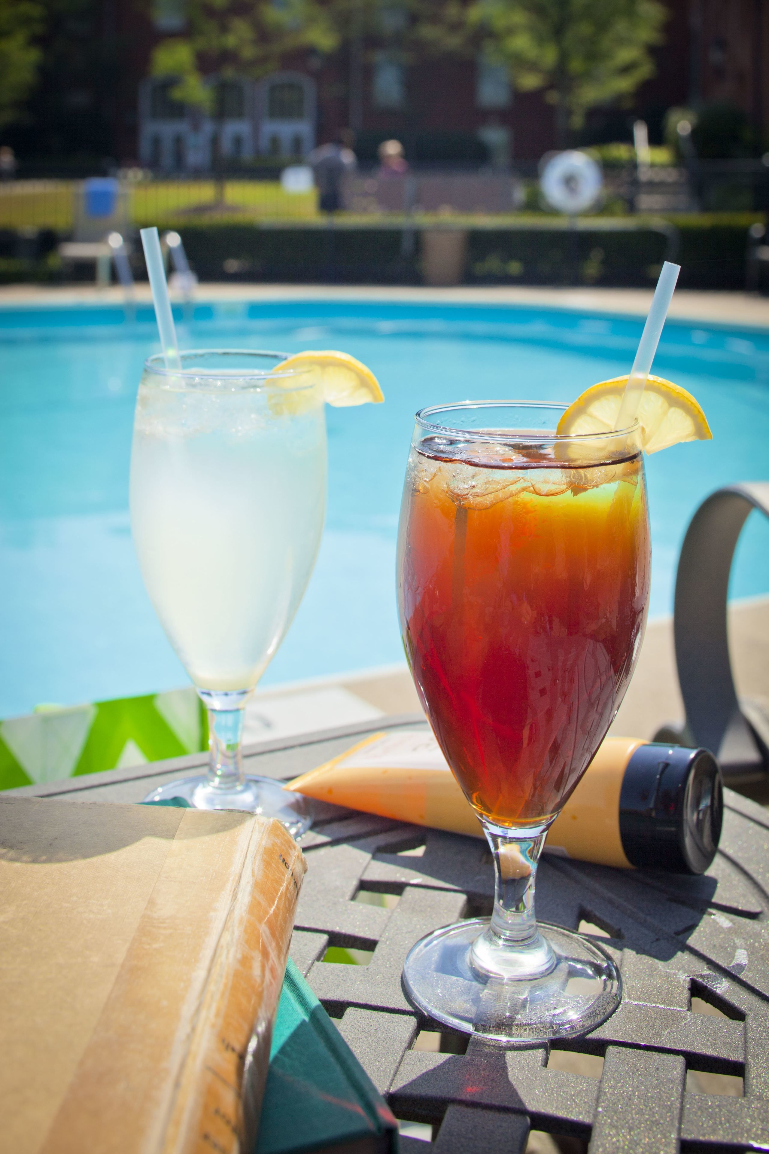 Dos bebidas sobre una mesa, cerca de una piscina, con una silla y una tabla de madera.