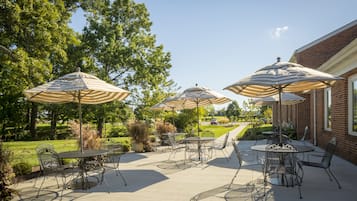 An outdoor patio with tables and chairs, surrounded by trees and a brick building.