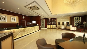 Un hall d’hôtel doté d’un comptoir de réception en marbre, d’une horloge, d’un lustre et de plusieurs espaces aménagés avec des sièges.