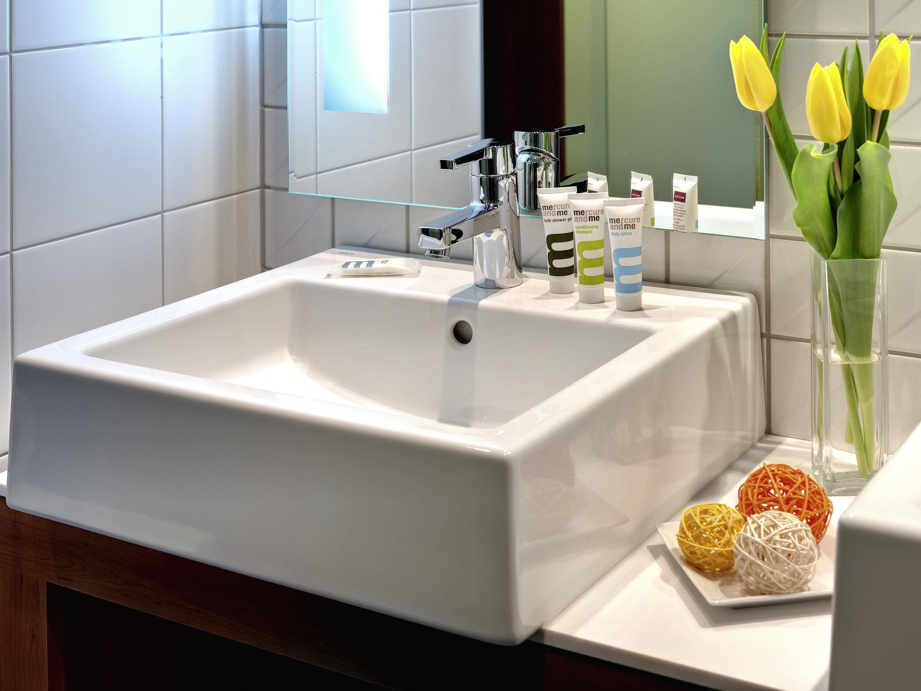 Un baño con un lavamanos blanco, grifo cromado y artículos de aseo. Hay tulipanes amarillos en un jarrón sobre la encimera.