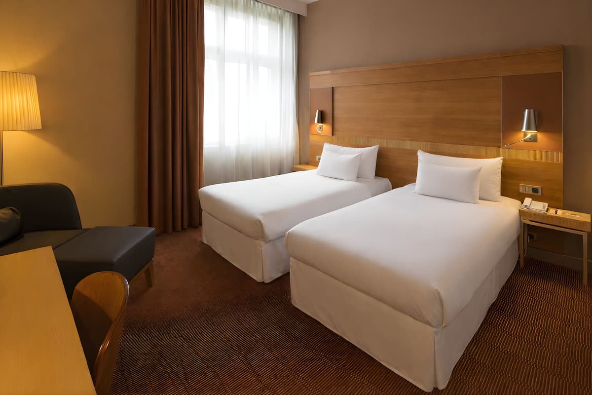 A hotel room with two beds, a desk, a chair, and a lamp.