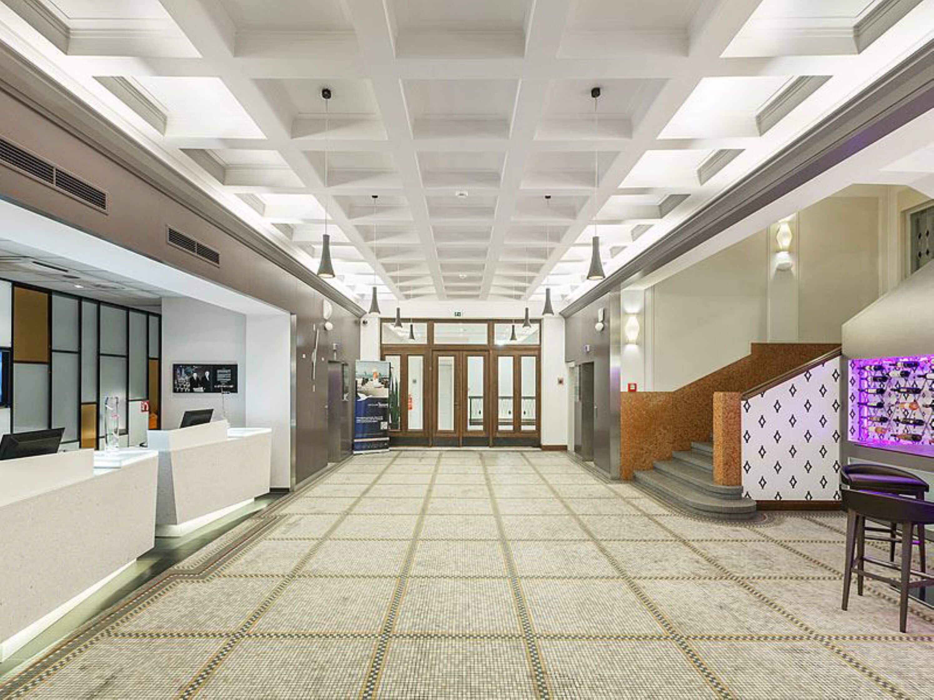 A spacious lobby with a high ceiling, a reception desk, a staircase, and a patterned floor.