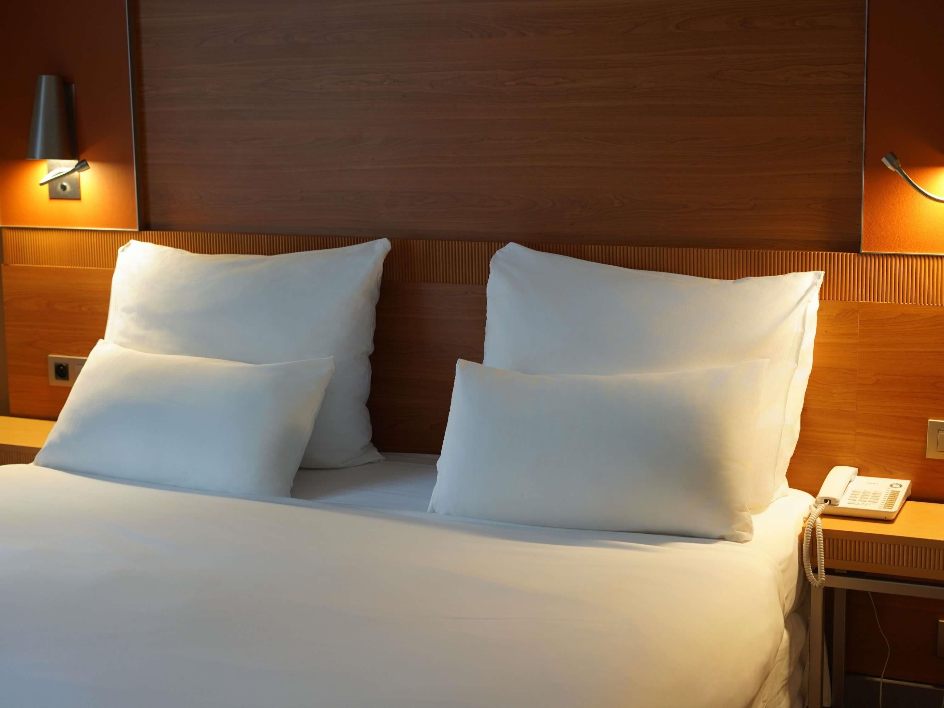 Une chambre d’hôtel avec une tête de lit en bois, deux oreillers blancs, une table de chevet avec un téléphone et des lampes fixées au mur.