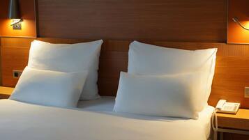 A hotel room with a wooden headboard, two white pillows, a bedside table with a phone, and wall-mounted lighting.