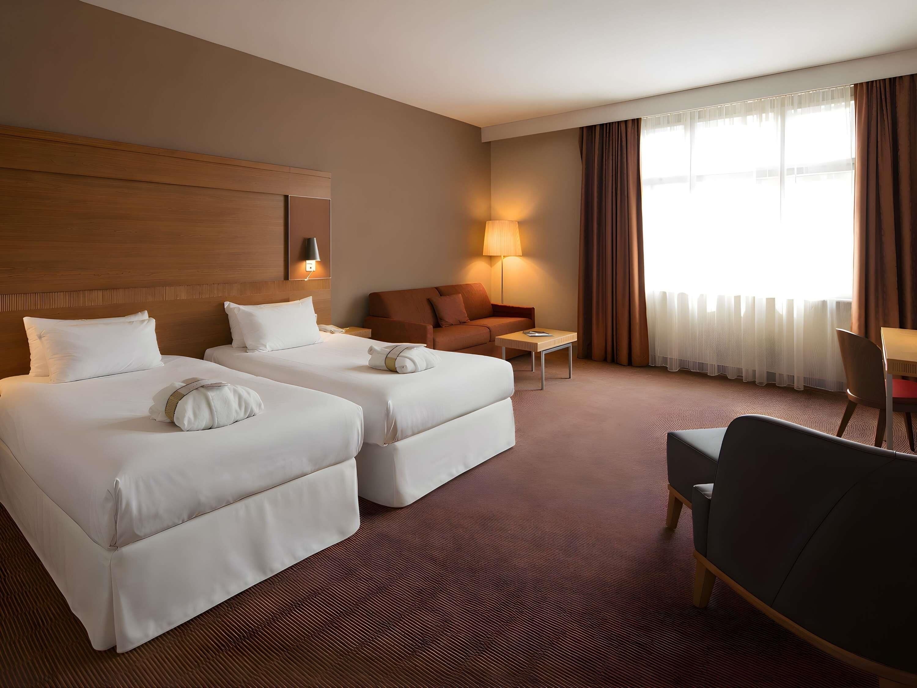 A hotel room with two beds, a sofa, a small table, a chair, and a lamp.