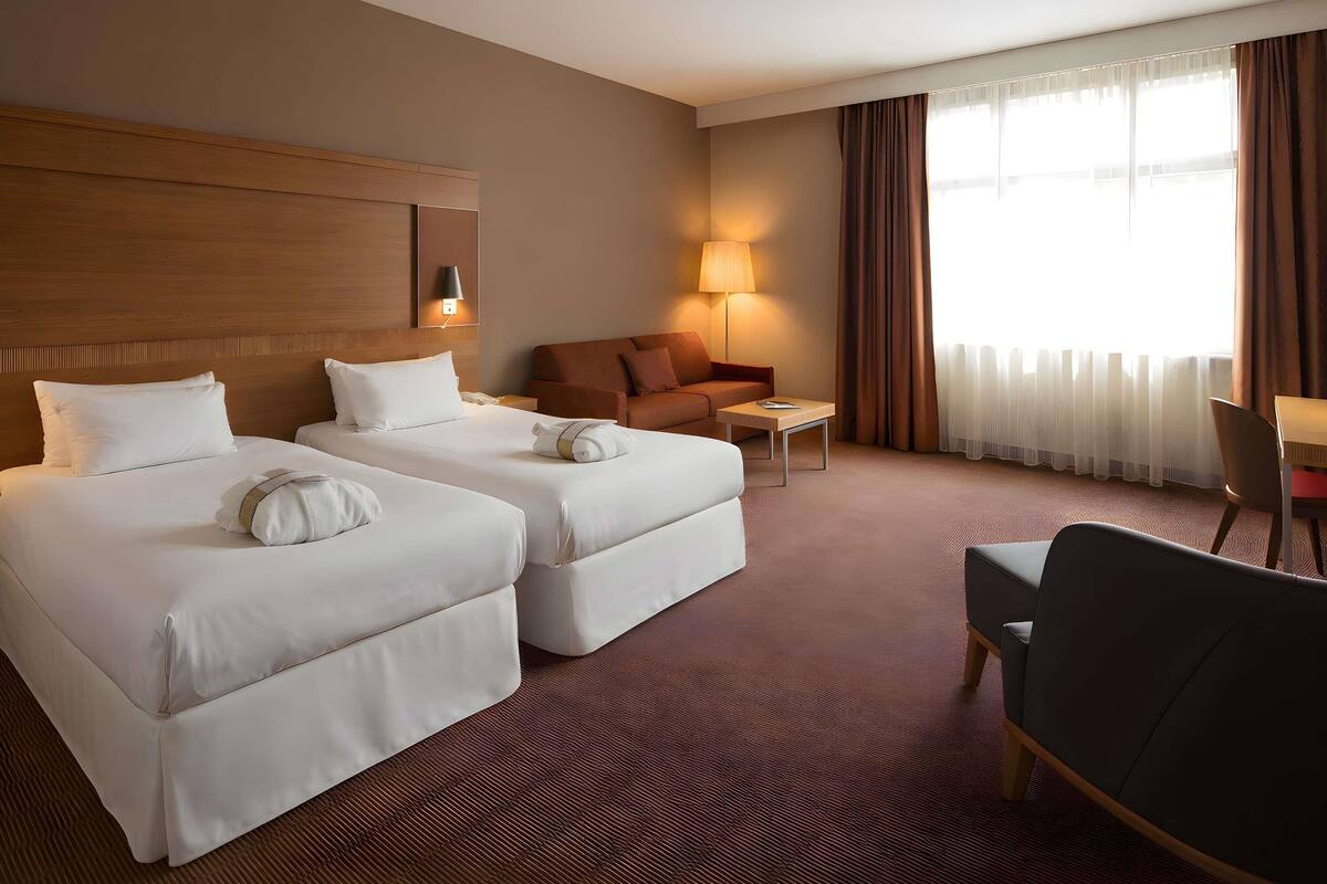 A hotel room with two beds, a sofa, a small table, a chair, and a lamp.