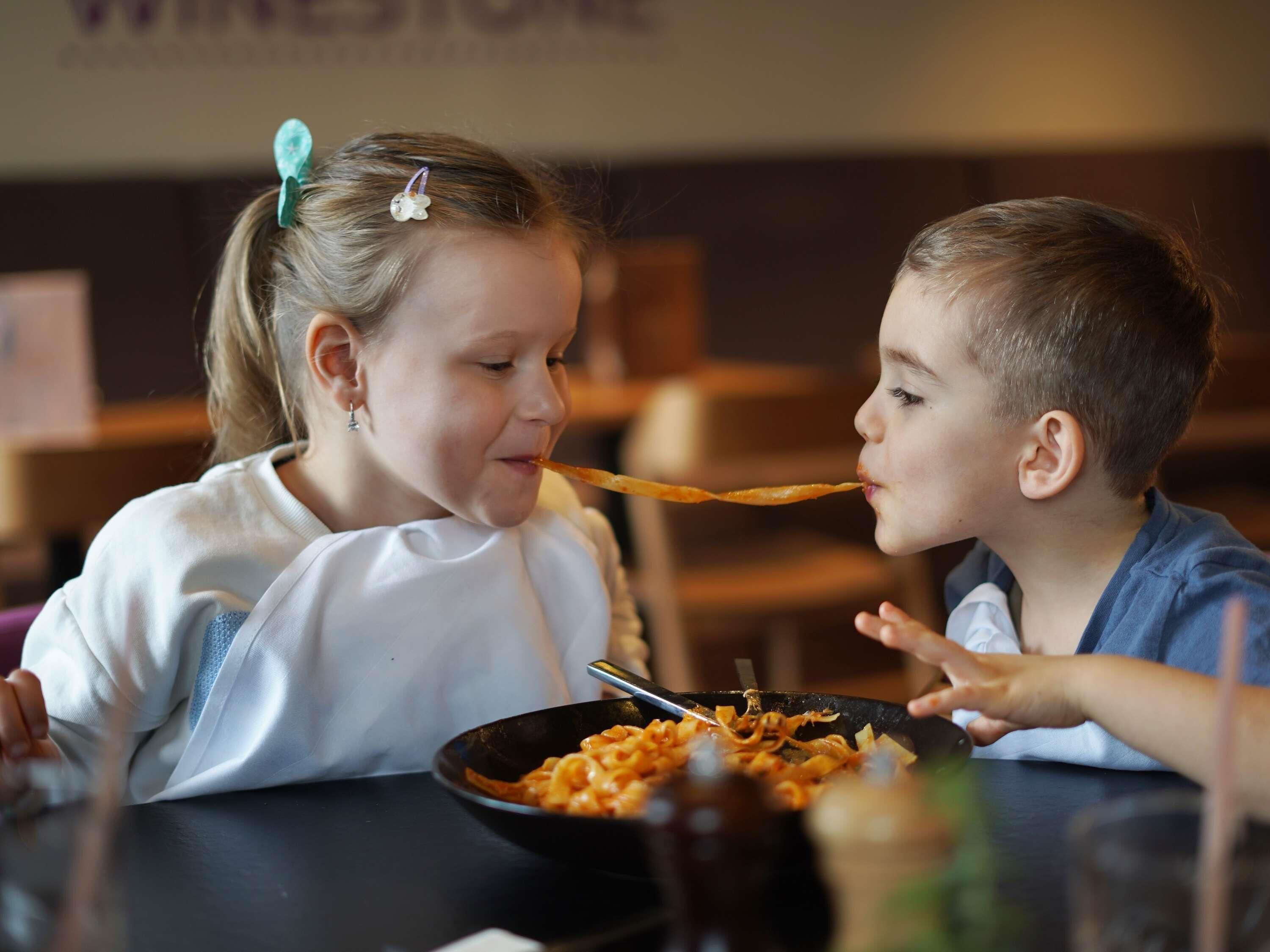 Two children eating at a table with a bowl of food in front of them.