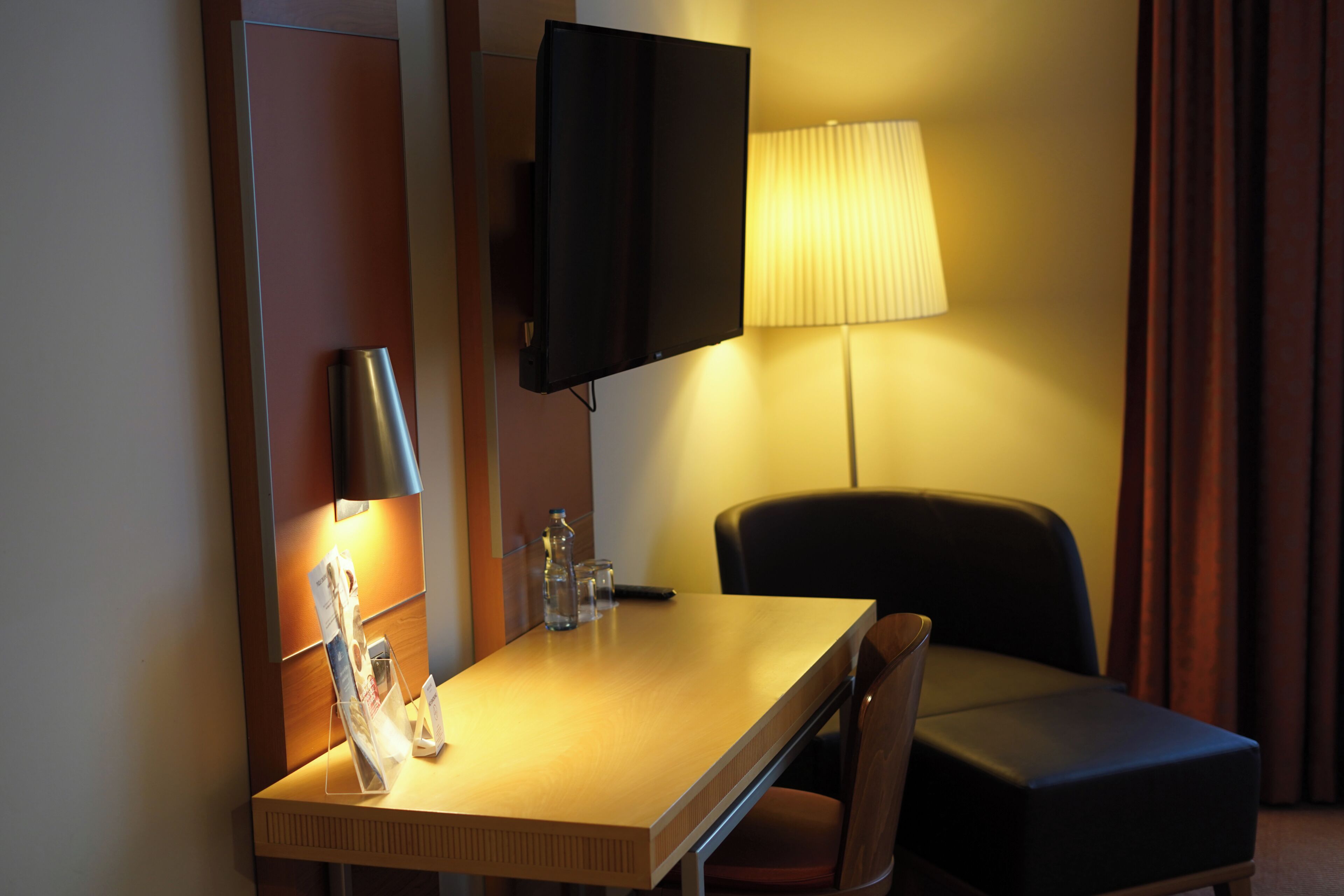A hotel room with a desk, a chair, a television mounted on the wall, a lamp, and a window with curtains.