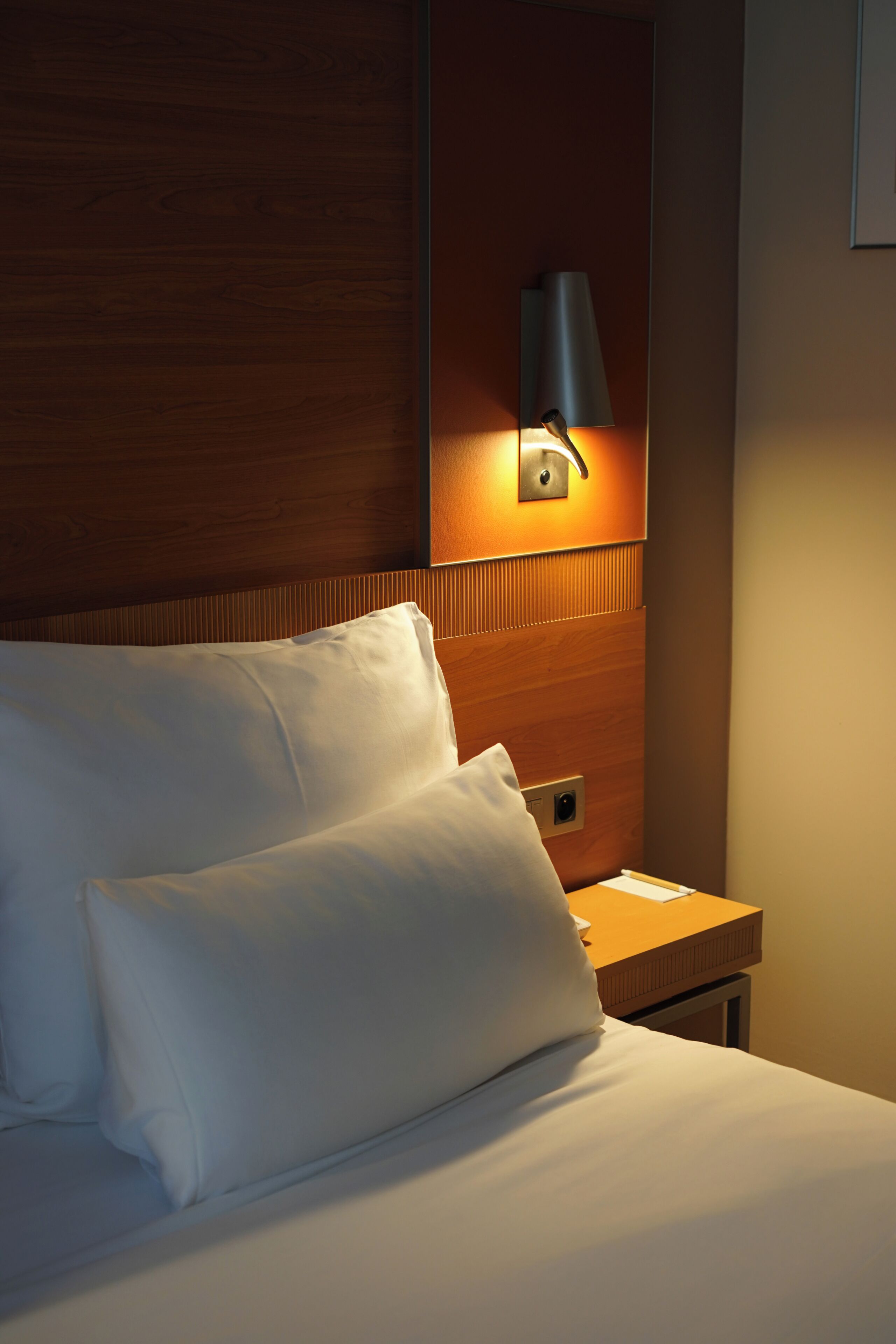 A hotel room with a wooden headboard, a bedside lamp, and a nightstand with a lamp.