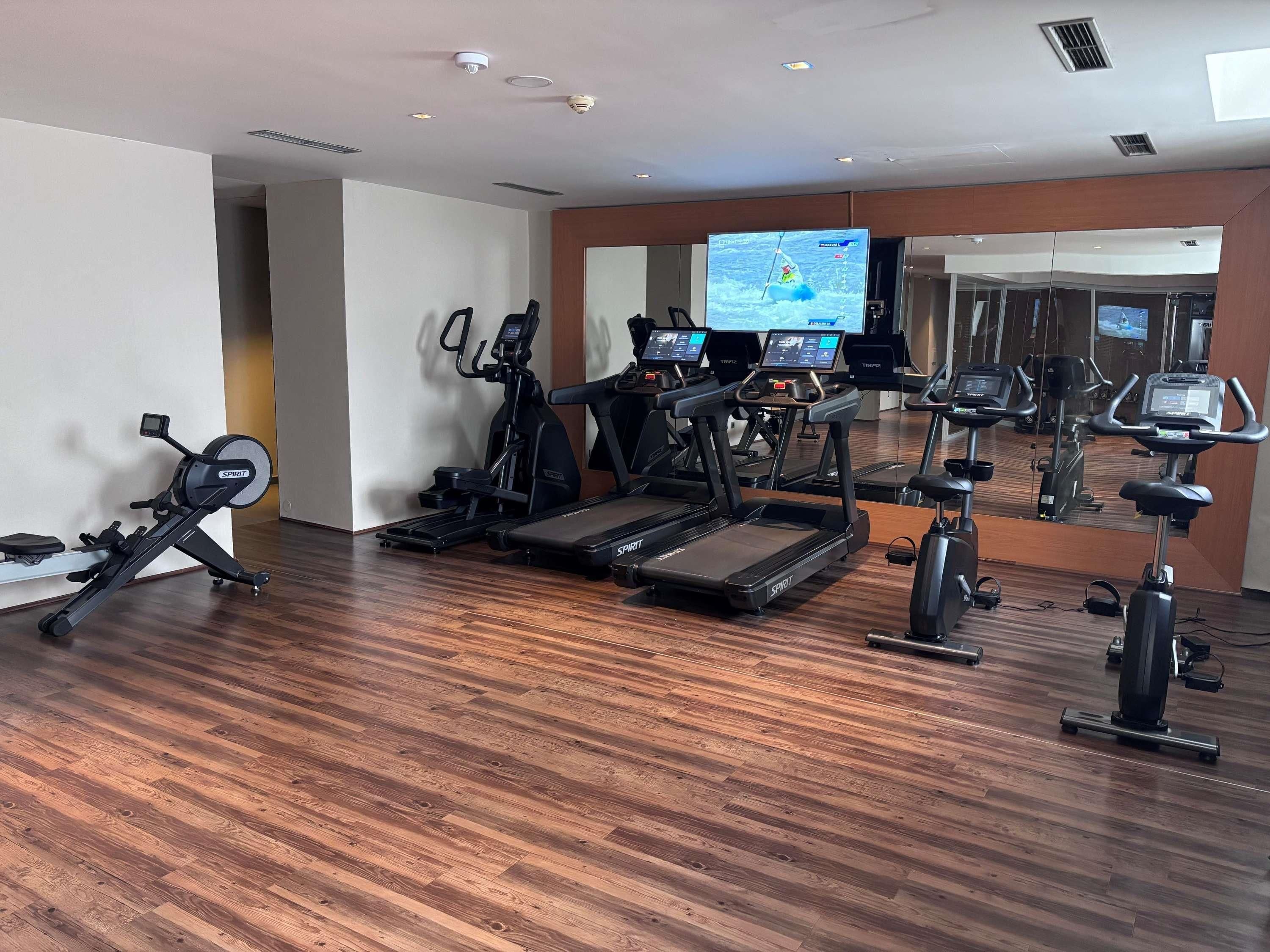 A gym with various exercise equipment, including treadmills and stationary bikes, and a television displaying a sports program.