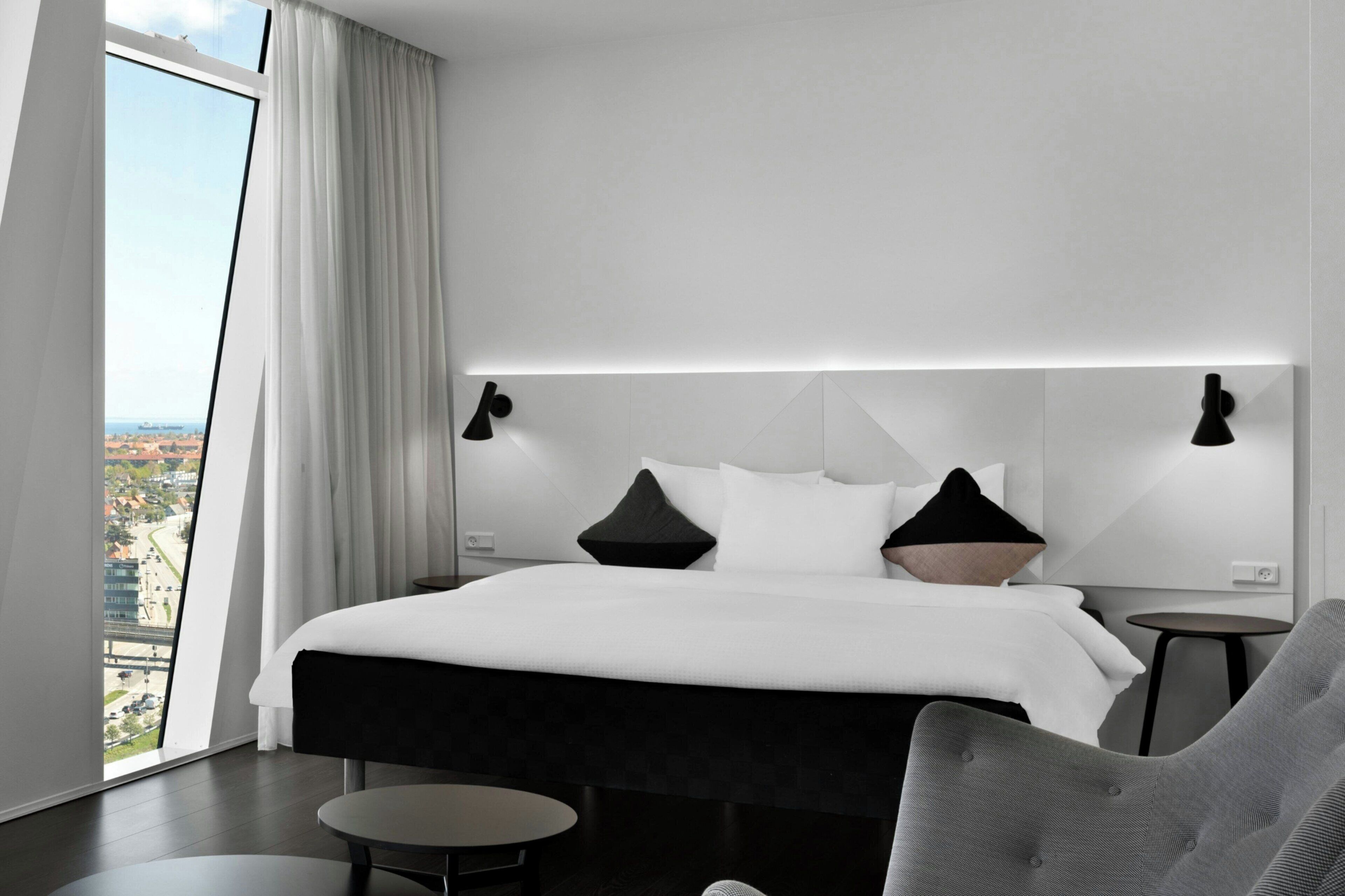 A modern hotel room with a large bed, two bedside tables, a chair, and a view of the city.