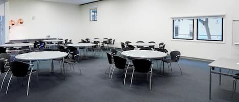 A conference room with multiple tables and chairs, white walls, and large windows.