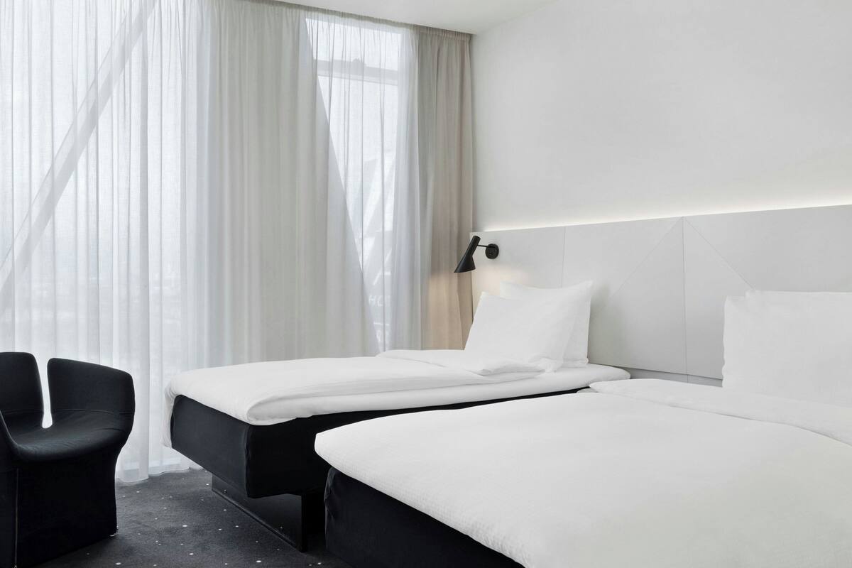 A hotel room with two beds, a black chair, and a large window with sheer curtains.