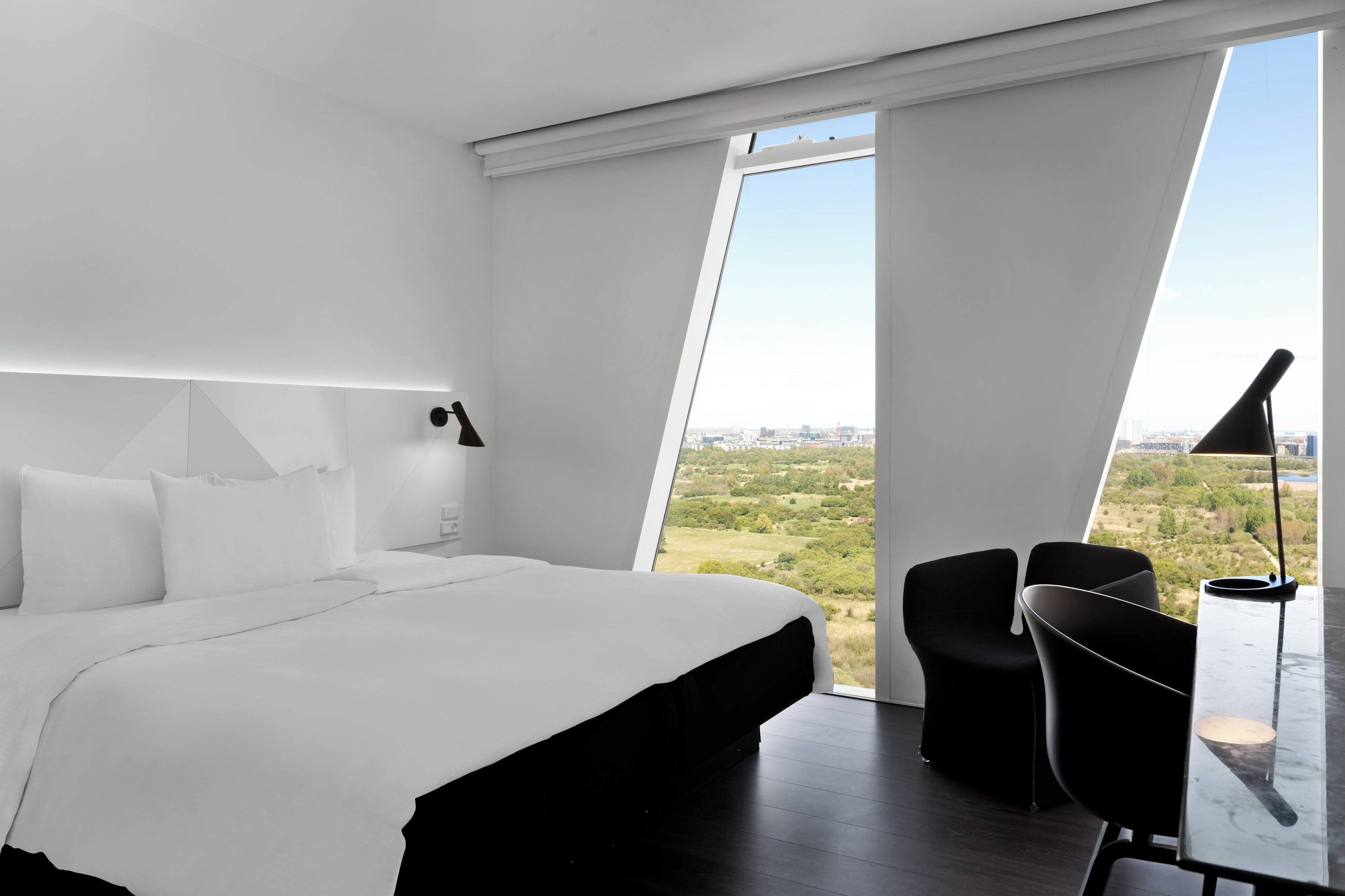 Una habitación de hotel moderna con una cama grande, un escritorio y vistas a un paisaje urbano.