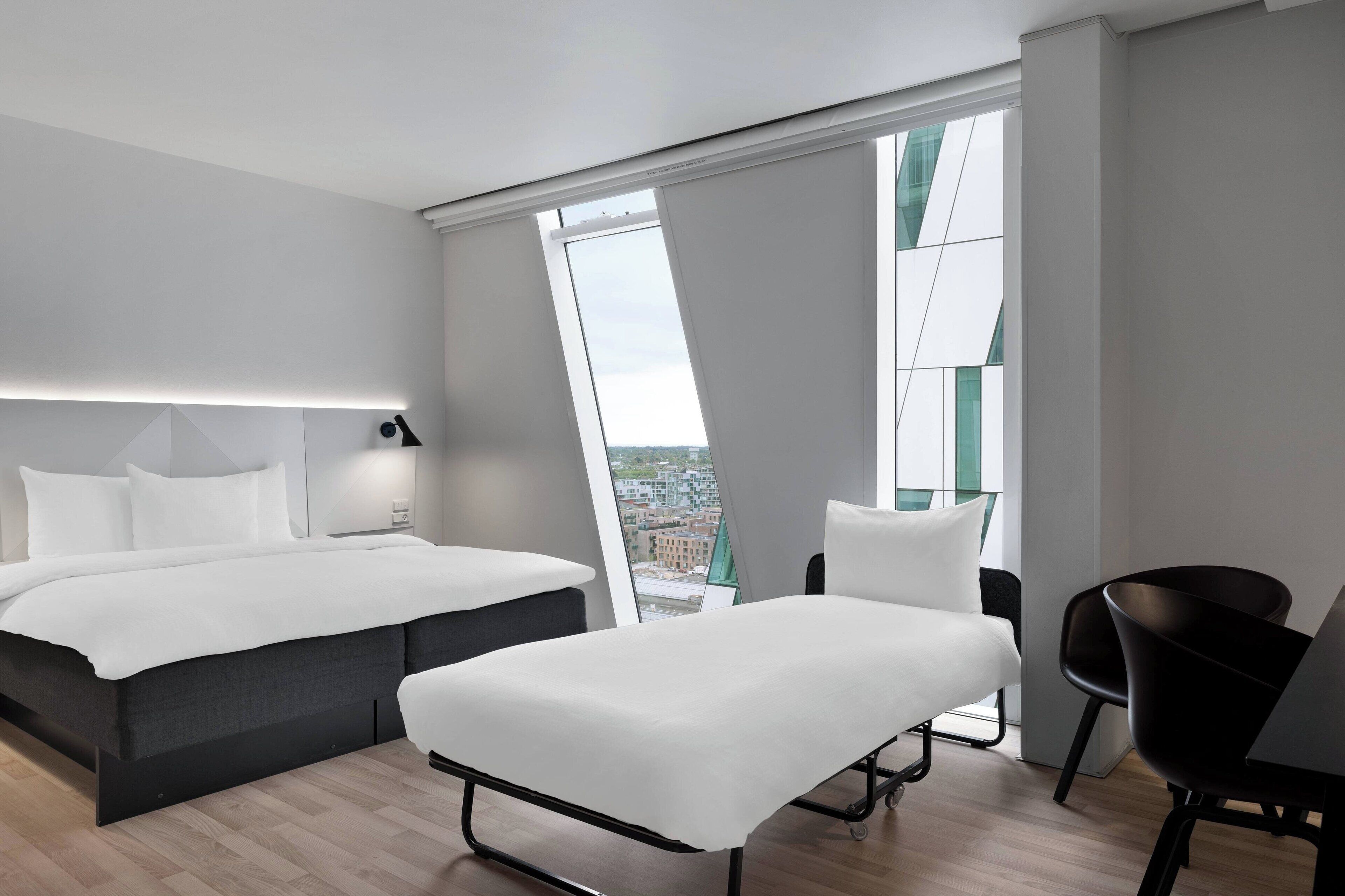 Quarto de hotel moderno com uma cama grande, mesa de cabeceira e vista da cidade através de janelas do chão ao teto.