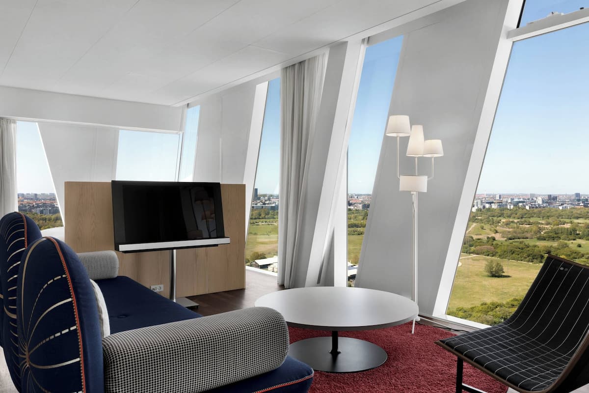 A modern hotel room with a large window, a flat-screen TV, a round table, and a sofa.