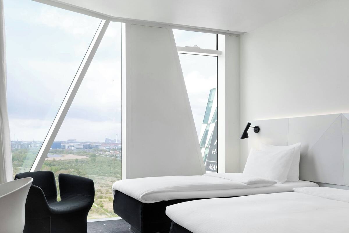 A modern hotel room with two beds, a large window offering a city view, and a black chair.