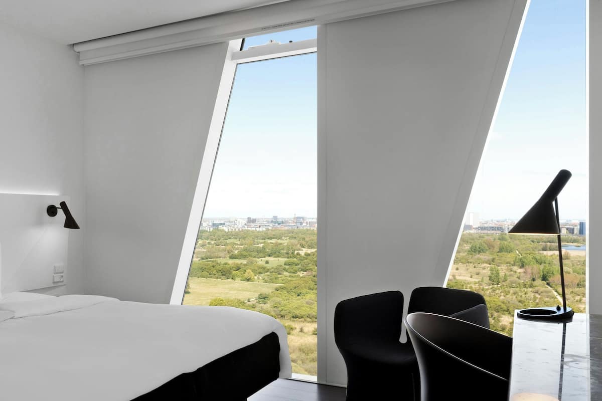 A modern hotel room with a large bed, a desk, and a view of a cityscape and greenery.