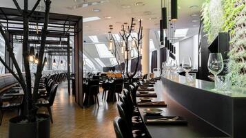 A modern restaurant interior with a long dining table, black leather chairs, and a bar area with a green wall.