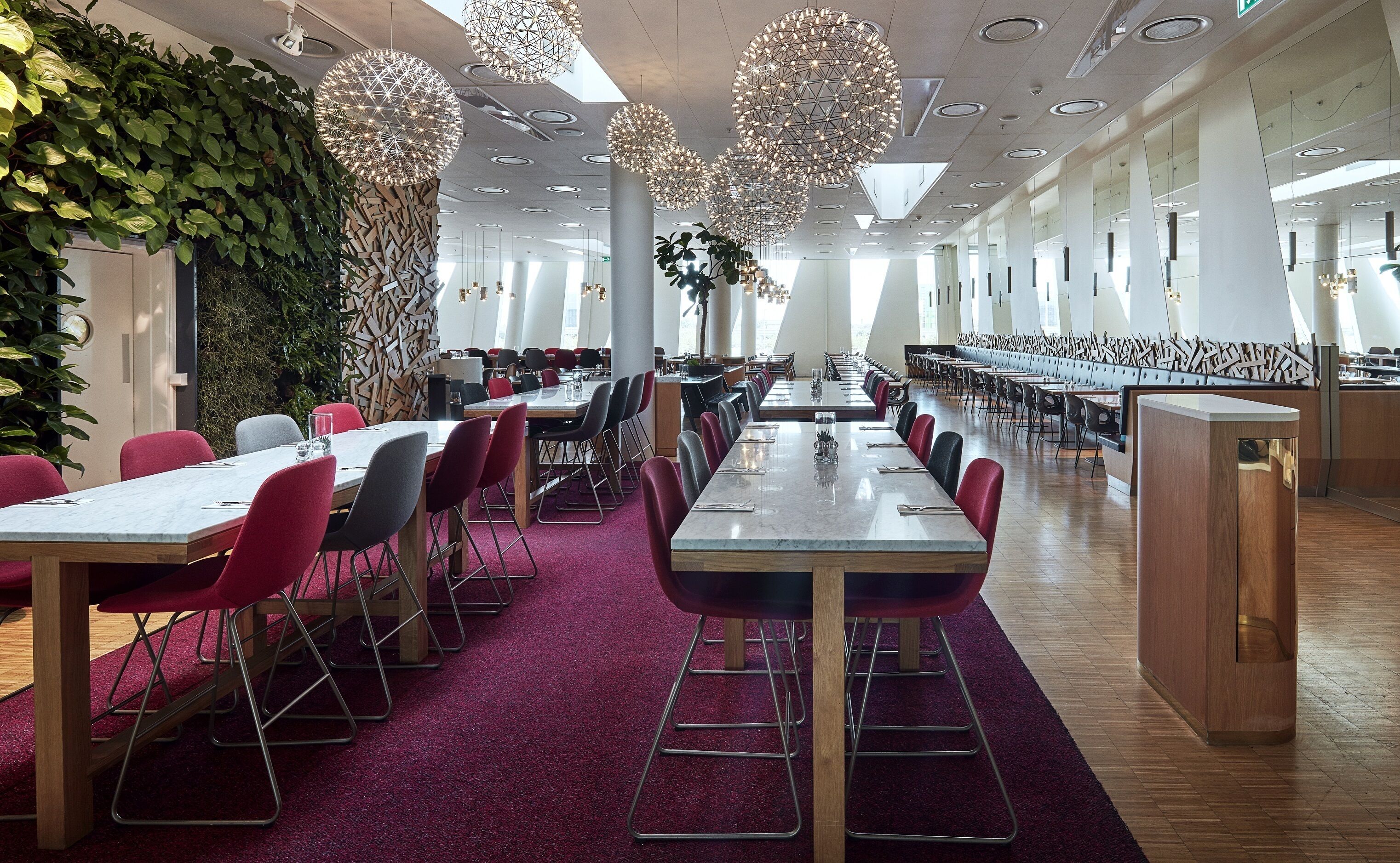 Uma sala de jantar moderna com mesas compridas, cadeiras vermelhas e pretas, e luminárias pendentes decorativas.