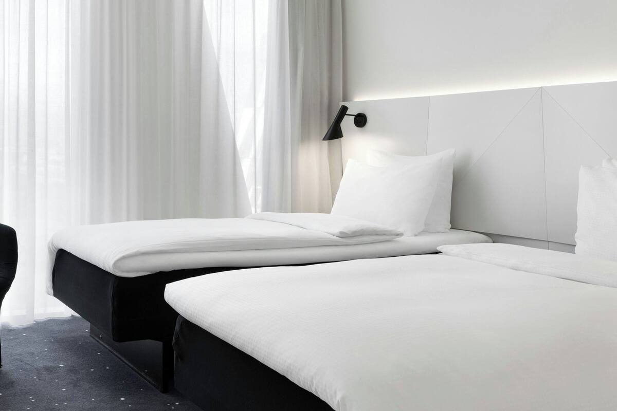 A modern hotel room with two single beds, a black nightstand, a wall-mounted lamp, and sheer curtains.
