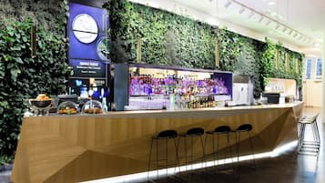 Een moderne bar met een houten toonbank, barstoelen en een groene wand met planten.
