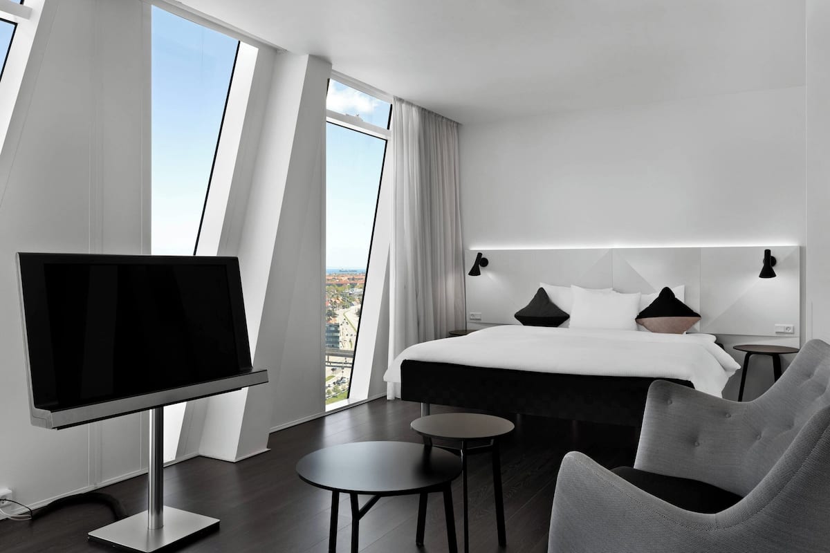 A modern hotel room with a large bed, a flat-screen TV, a small table, and a comfortable chair.