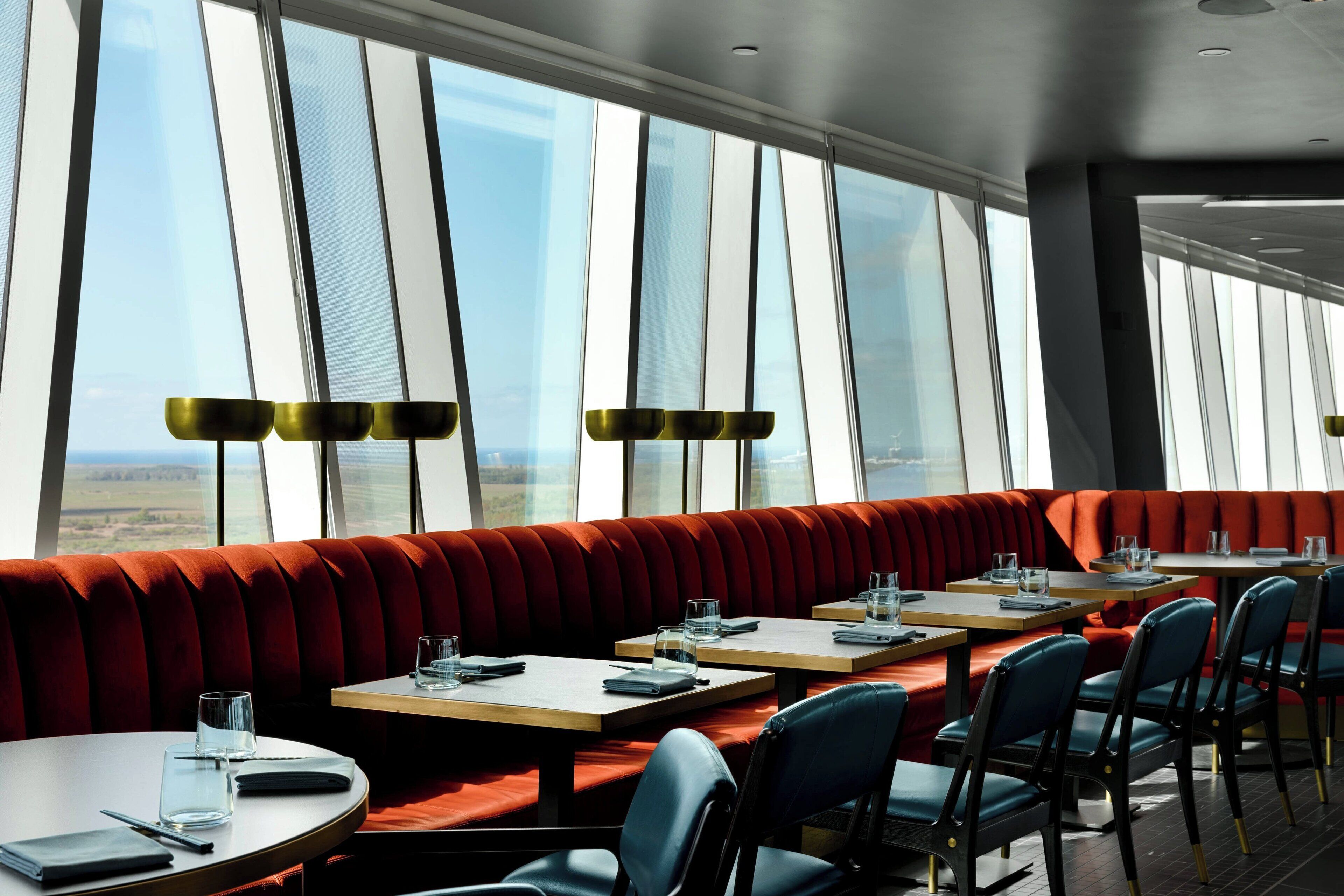 A restaurant with a long red cushioned bench, tables with chairs, and large windows offering a view of the landscape.