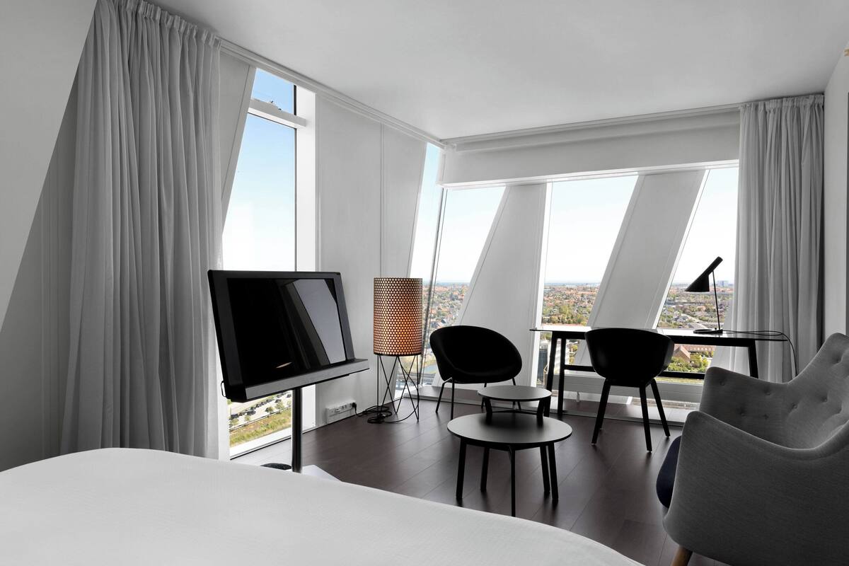 A modern hotel room with a large bed, a desk with a computer, two chairs, and a TV.