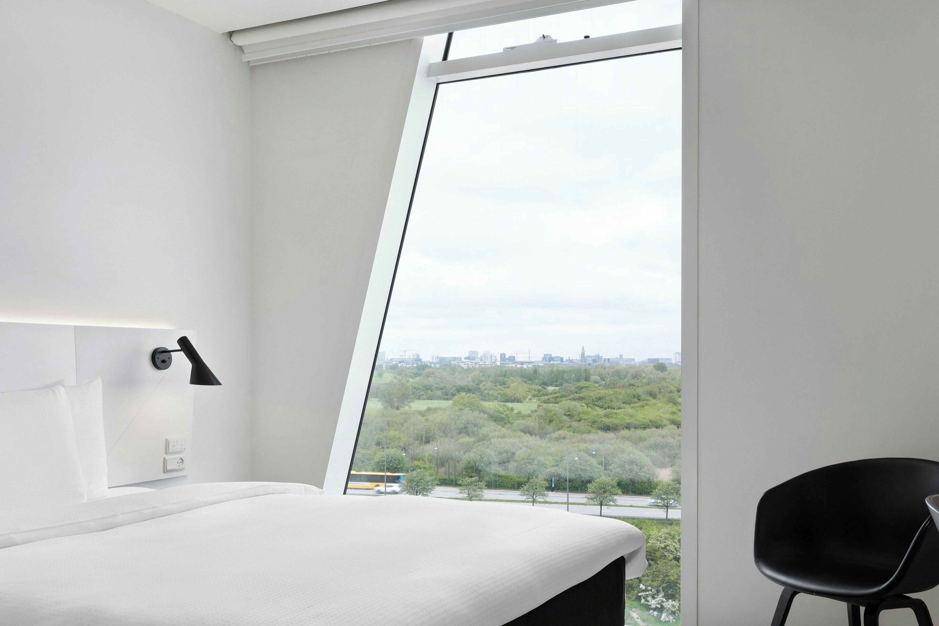 Een moderne hotelkamer met een groot raam, een bed, een zwarte stoel en een wandlamp.
