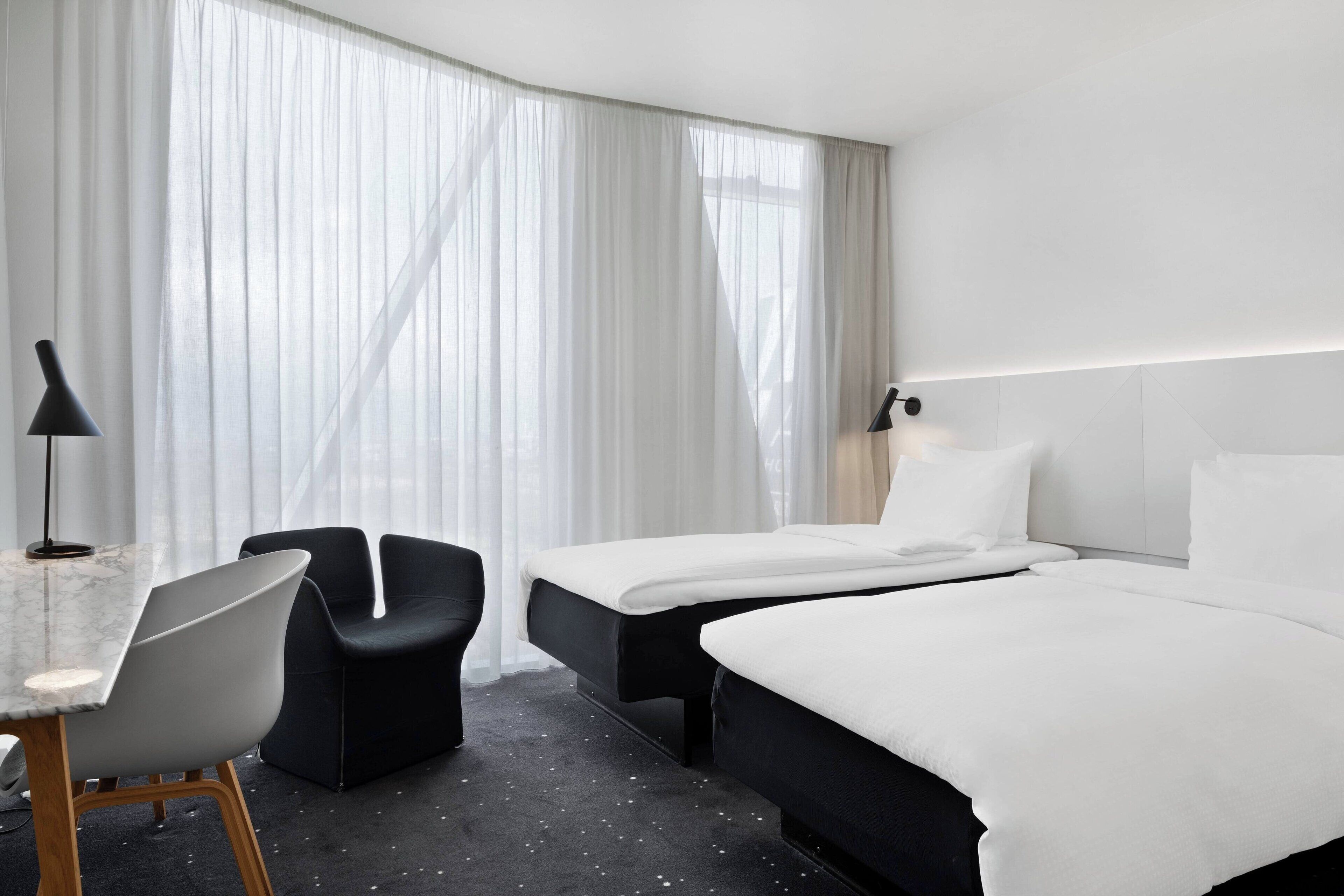 A hotel room with a bed, two bedside tables, a desk with a chair, and a large window with sheer curtains.