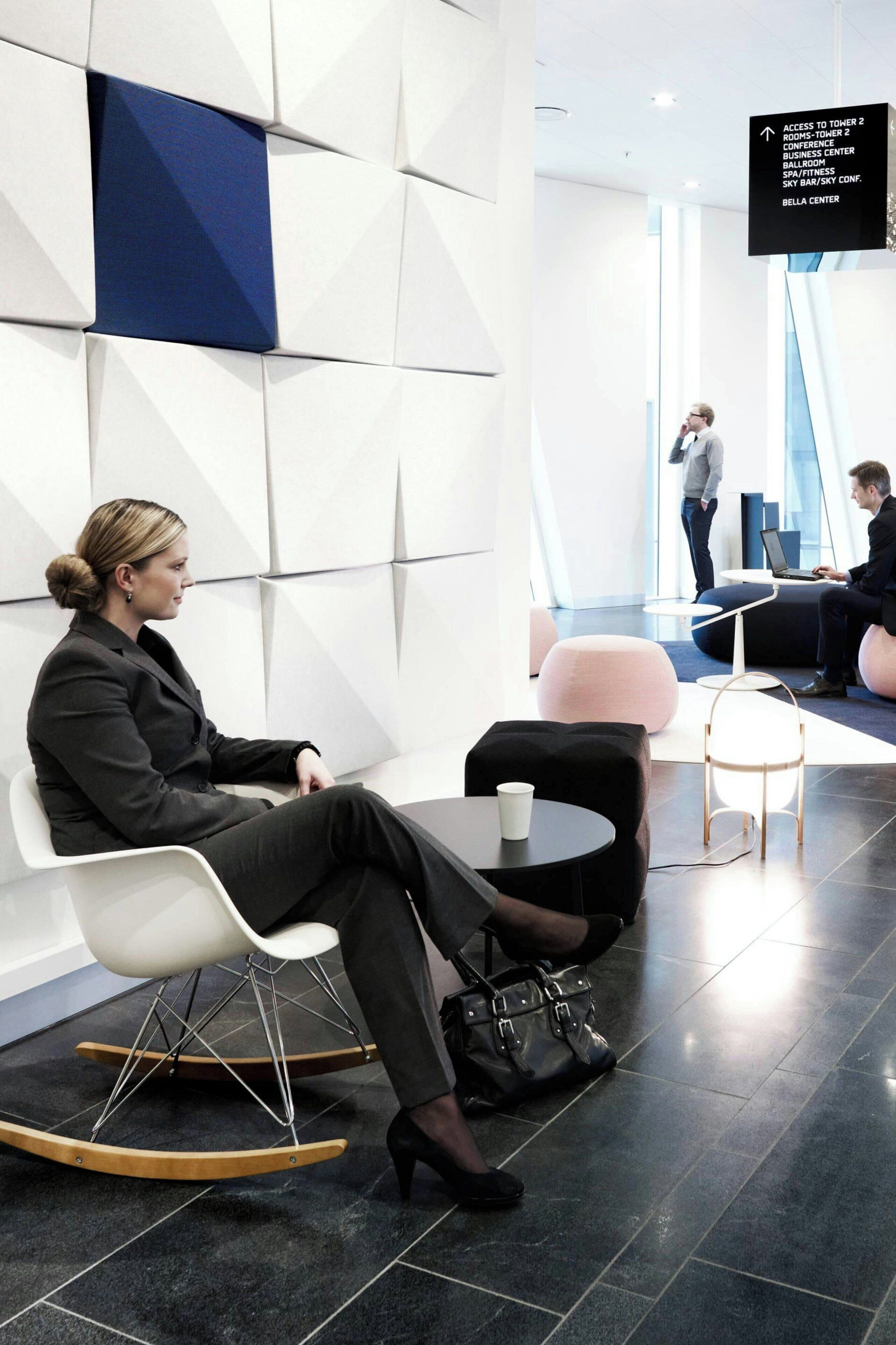 A modern interior with a woman seated at a table, a man working on a laptop, and another person standing.