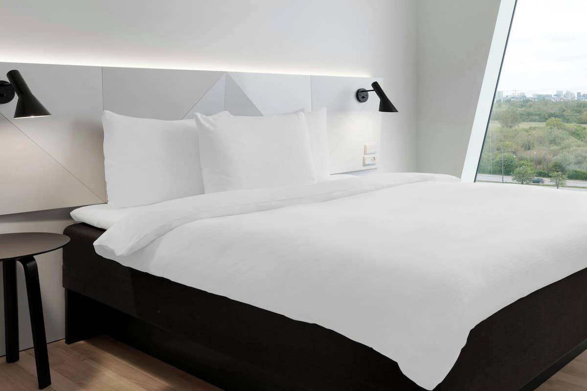 A modern hotel room with a large bed, two bedside lamps, a small round table, and a view of greenery through a large window.