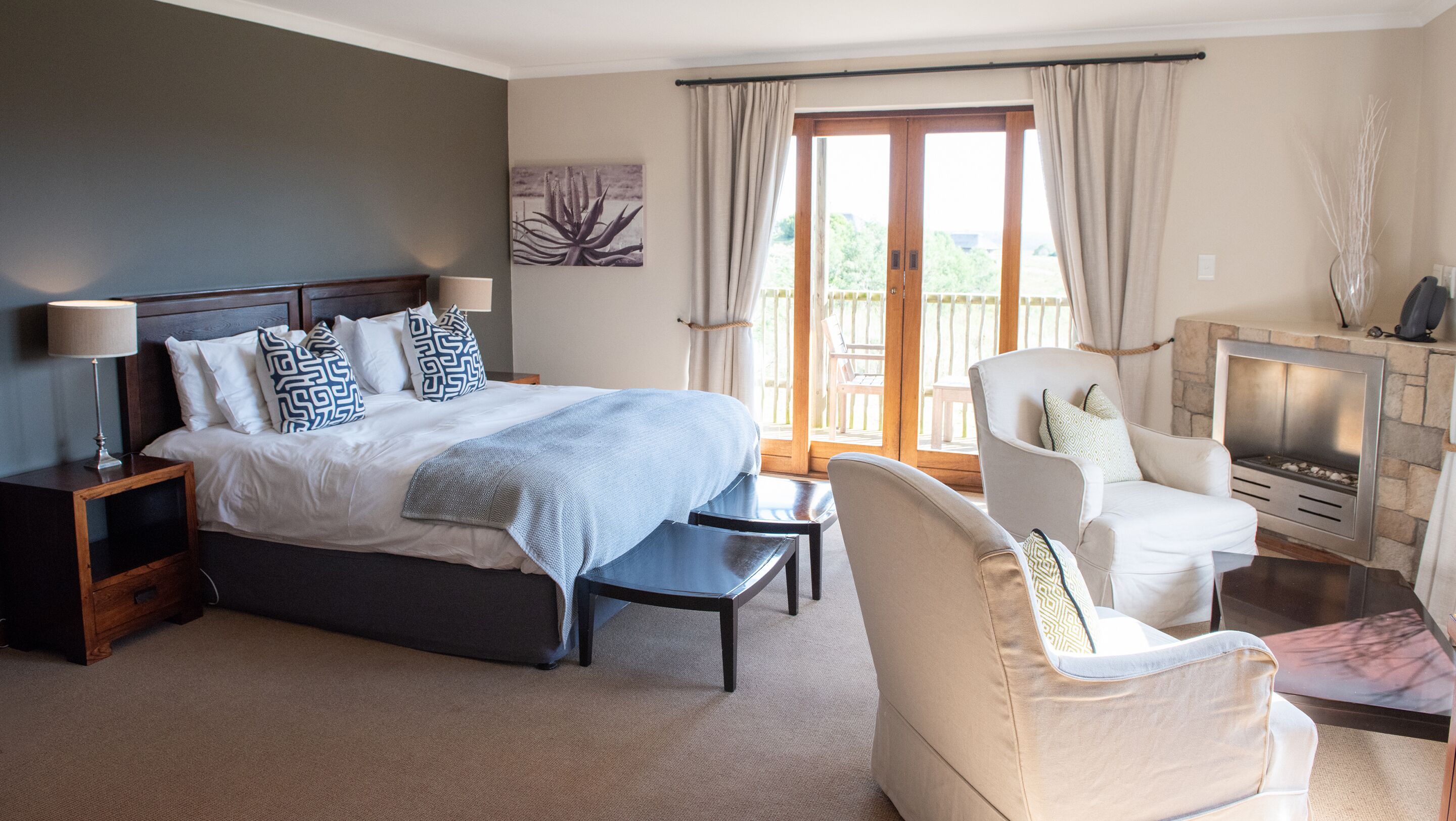 A hotel room with a bed, a chair, a small table, a fireplace, and a balcony with curtains.