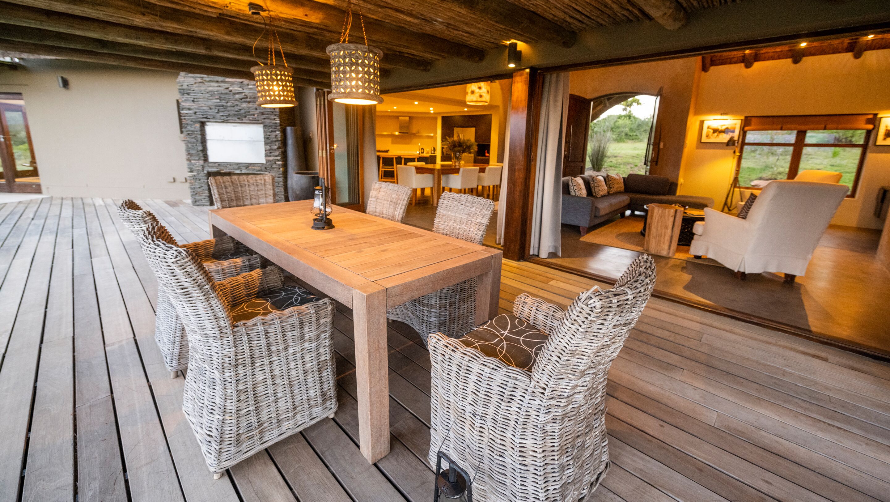 Una terraza de madera con mobiliario de mimbre, una mesa de comedor y luces colgantes.