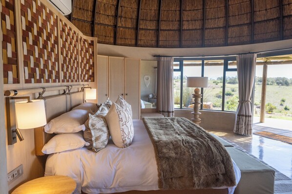 A spacious bedroom with a large bed, multiple pillows, a headboard with a woven pattern, a bedside table with a lamp, and a view of the outdoors through large windows. - Gondwana Game Reserve (Mossel Bay)