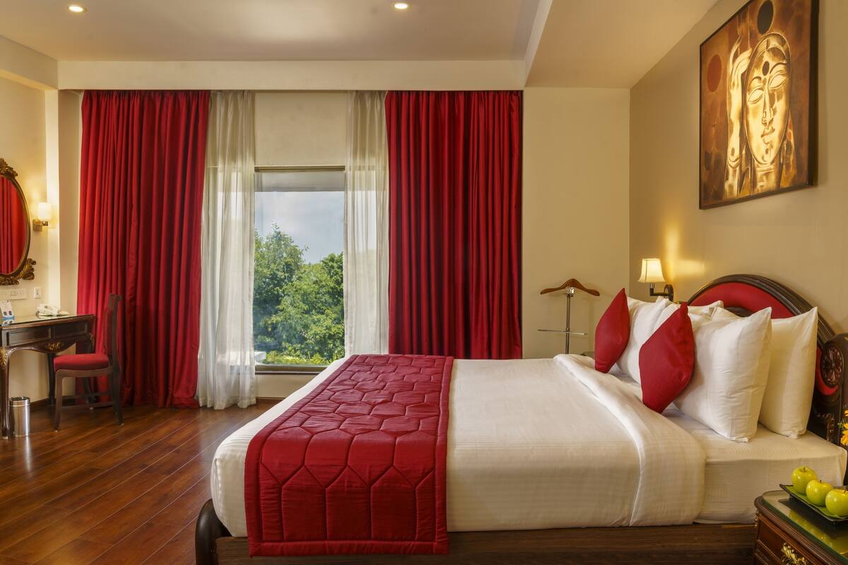 Een hotelkamer met een groot bed, rode gordijnen, een bureau met telefoon en een schilderij aan de muur.