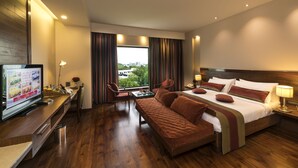 A hotel room with a large bed, a sofa, a desk with a TV, and a window with curtains.