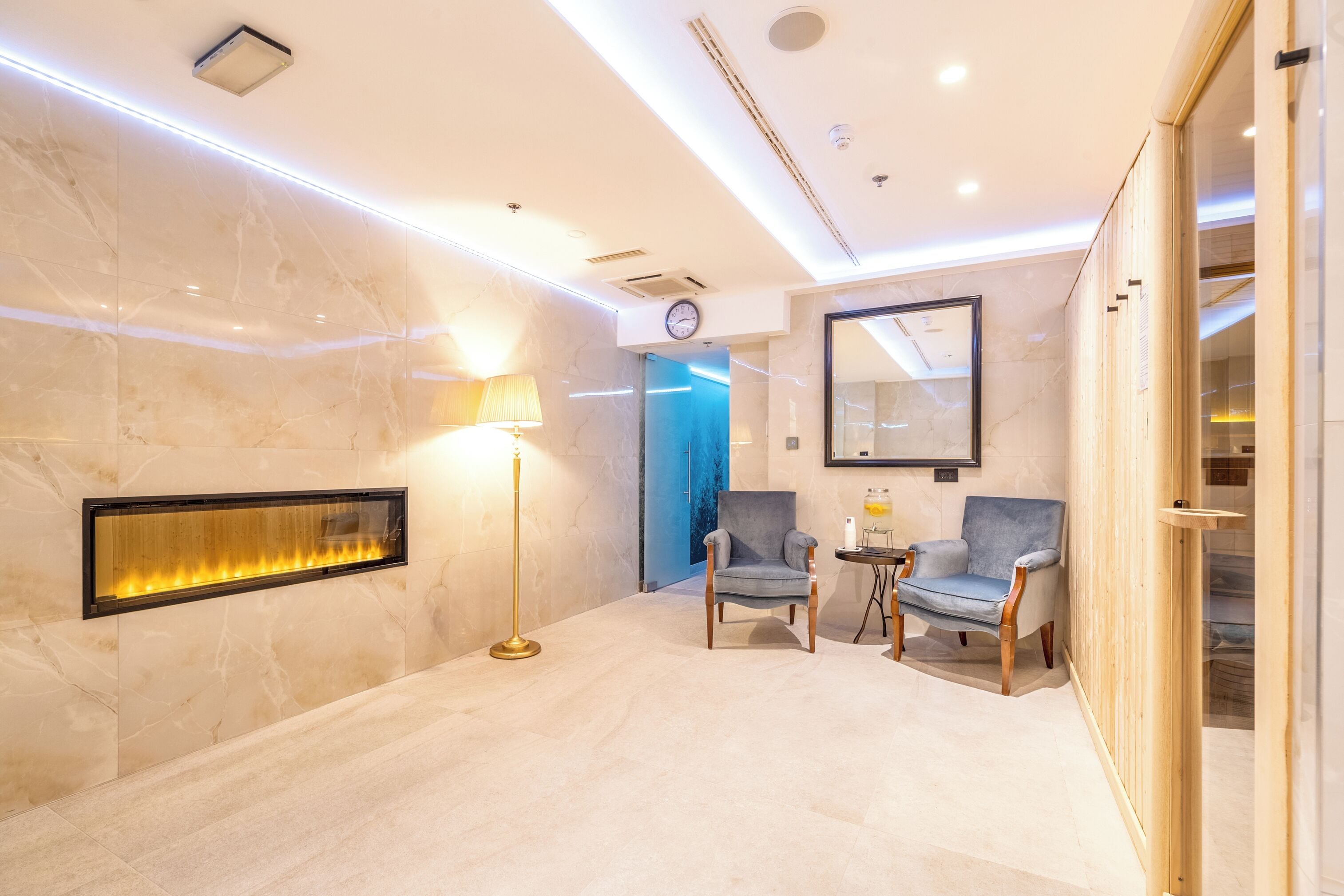 A modern lobby with marble flooring, a wall-mounted fire feature, two armchairs, a floor lamp, and a clock on the wall.
