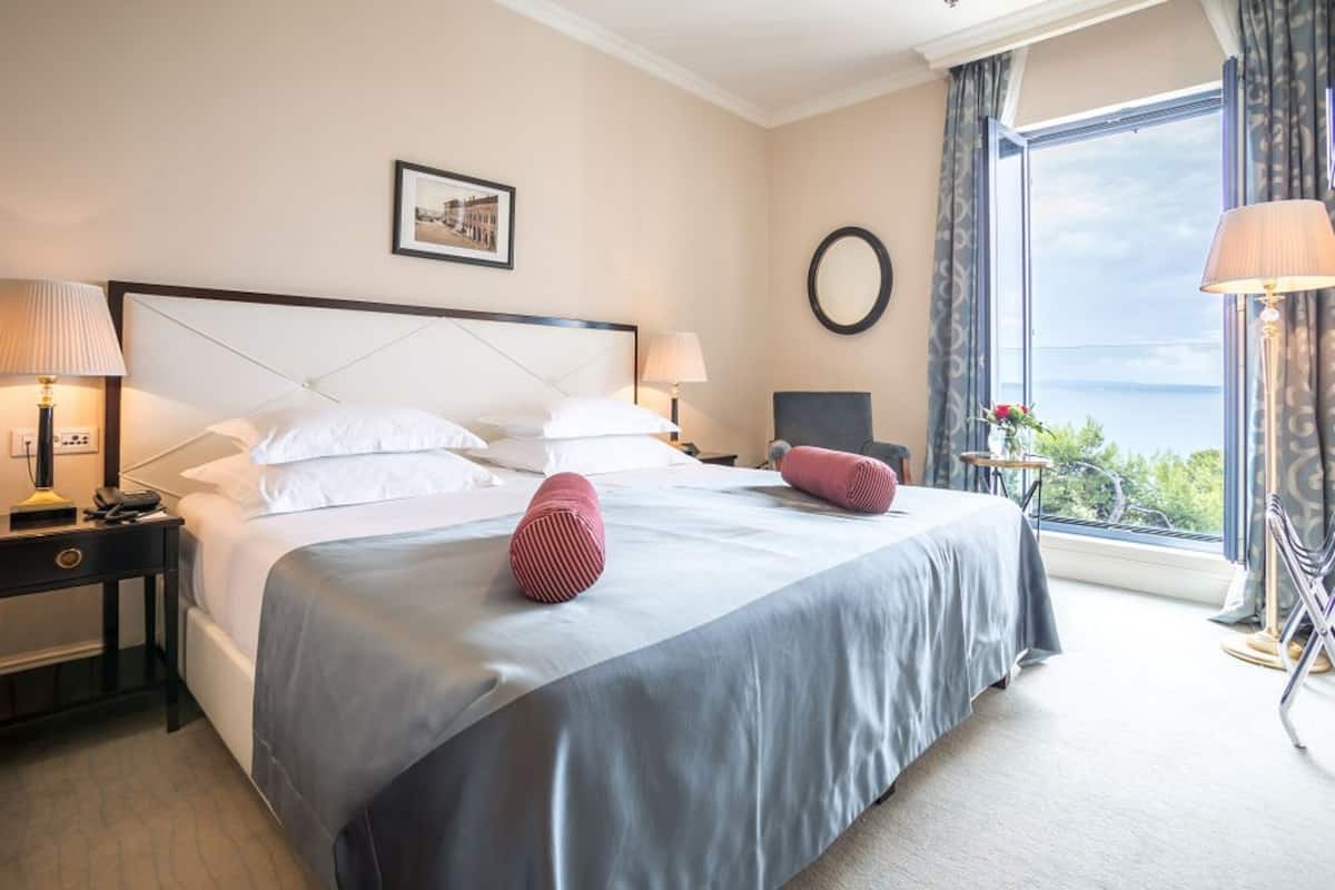 A hotel room with a large bed, two bedside lamps, a chair, a small table, and a view of the sea.