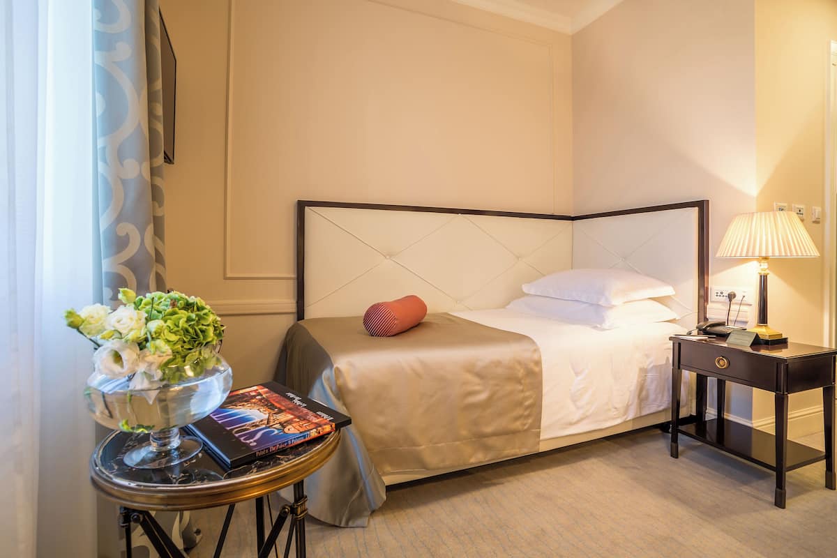 A neatly arranged hotel room with a bed, bedside table, lamp, and a small table with a vase of flowers and books.