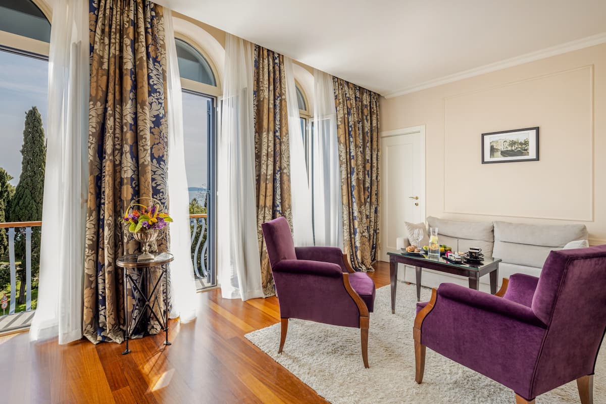 A spacious living room with a large window, a sofa, two purple armchairs, and a small table with a vase of flowers.