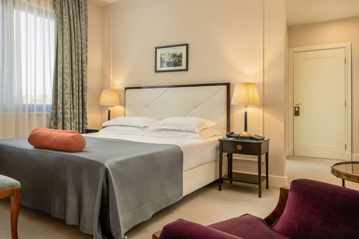 A hotel room with a bed, two bedside tables with lamps, a chair, a door, and a framed picture on the wall.