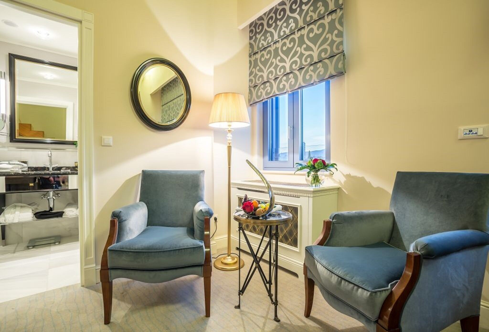 A living room with two blue armchairs, a floor lamp, a round mirror, and a window with a patterned blind.