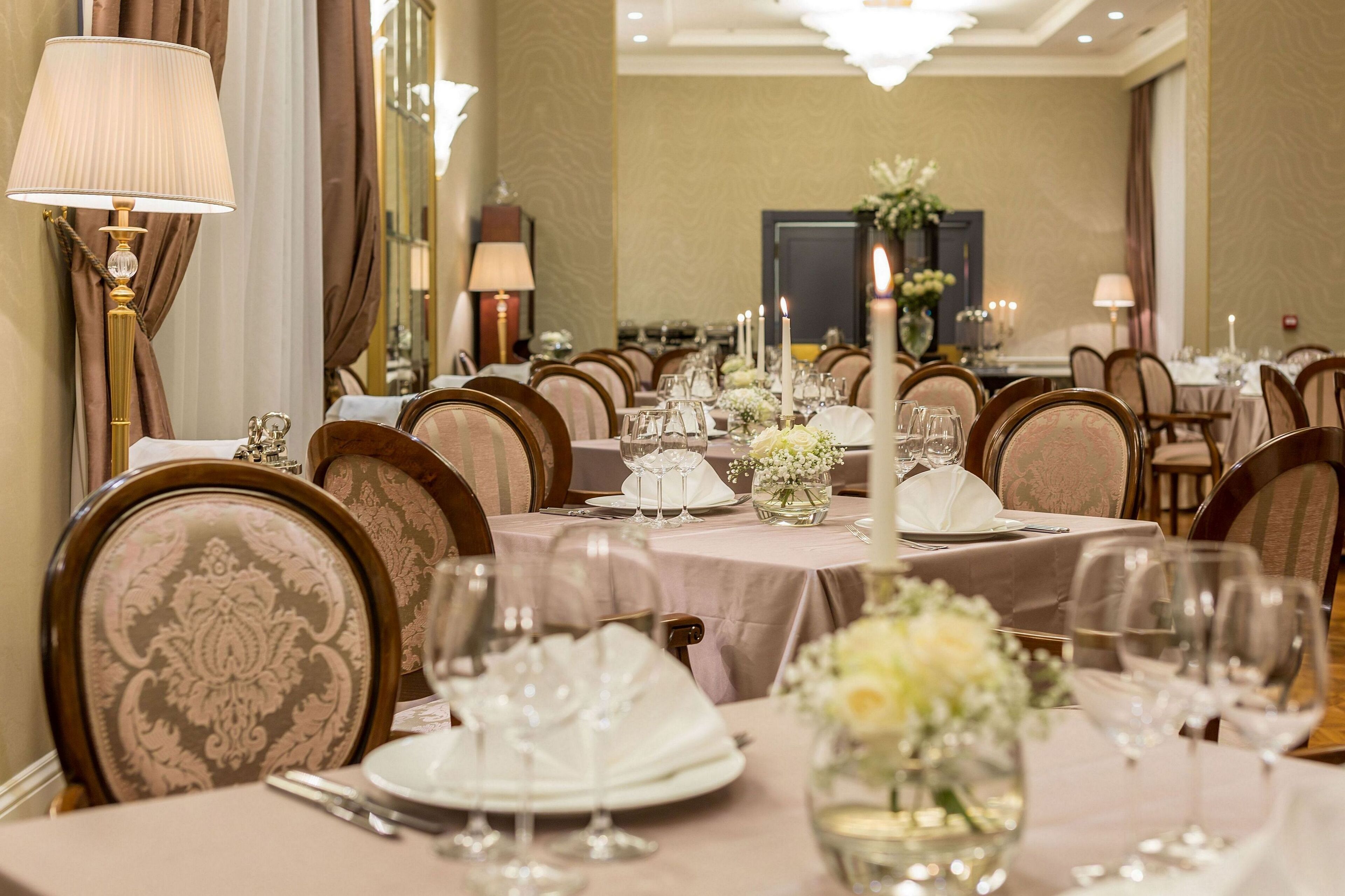 A formal dining room with round tables, chairs, and elegant table settings including white tablecloths, glassware, and floral centerpieces.