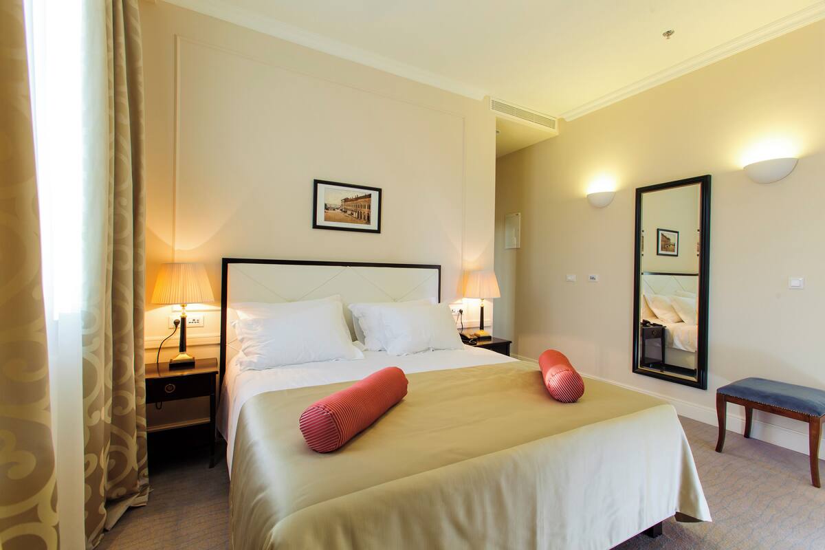 A hotel room with a large bed, two bedside tables with lamps, a bench, a mirror, and a picture on the wall.