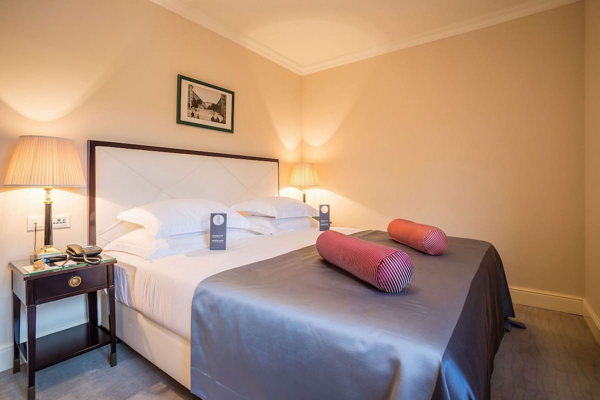 A hotel room with a bed, two bedside tables with lamps, a framed picture on the wall, and a red pillow on the bed.